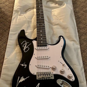 Puede incluir: Guitarra eléctrica negra con golpeador blanco y firmas. La guitarra tiene mástil y pala de madera con la marca "Sclary". El cuerpo es negro con detalles blancos y herrajes cromados.