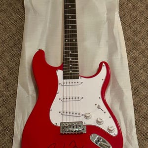 Könnte beinhalten: Eine rote E-Gitarre mit weißem Schlagbrett und verchromten Hardware. Die Gitarre ist signiert mit "Toby Fox".