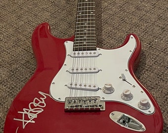 May include: A red electric guitar with a white pickguard and three pickups. The guitar has a signature in white on the body. It features a tremolo bridge, volume and tone knobs, and a maple neck.
