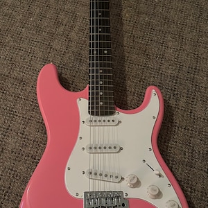 Könnte beinhalten: Eine rosa E-Gitarre mit weißem Schlagbrett und verchromten Hardware. Die Gitarre hat eine Signatur auf dem Korpus, die "JB" lautet.