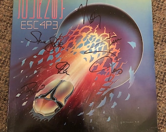 May include: A vinyl record album cover for Journey's Escape album. The cover features a spaceship flying through a field of broken glass and a large blue sphere in the background. The album cover is signed by all five members of Journey: Steve Perry, Neal Schon, Jonathan Cain, Ross Valory, and Steve Smith.