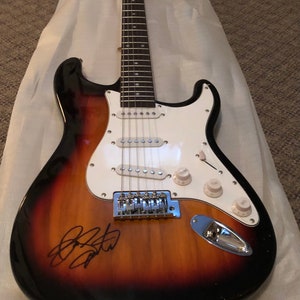 Może przedstawiać: Elektryczna gitara o wykończeniu sunburst z białym pickguardem i czarnym podpisem na korpusie. Gitara ma sześć strun i chromowany mostek.