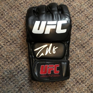 May include: Black UFC MMA training gloves with white UFC logo and red UFC branding. The gloves are signed in white ink.