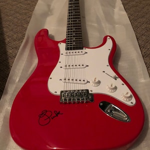 Könnte beinhalten: Eine rote E-Gitarre mit weißem Schlagbrett und verchromten Hardware. Die Gitarre ist mit "Pink" in schwarzer Tinte signiert.