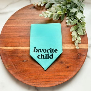 May include: A turquoise pet bandana with the text "favorite child" in black. The bandana is folded and placed on a round, wooden serving board. Greenery is arranged on the board.