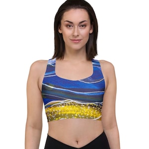 May include: A sports bra featuring a vibrant abstract print with deep blue, white, and yellow colors. The design includes wavy horizontal lines in the upper section and a textured, dotted yellow pattern across the lower portion of the garment.