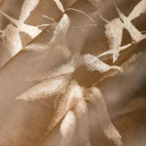 Puede incluir: Primer plano de una tela doblada con una superficie texturizada. La tela es de color marr&oacute;n c&aacute;lido con un patr&oacute;n de dise&ntilde;os blancos similares a pinceladas. La luz se refleja en la tela, creando reflejos y sombras.