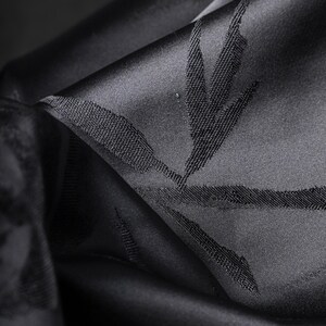 Puede incluir: Primer plano de tela negra con un brillo sutil. La tela est&aacute; doblada, revelando un patr&oacute;n texturizado de hojas o ramas. El color oscuro y el dise&ntilde;o detallado sugieren un textil lujoso, posiblemente para ropa o decoraci&oacute;n del hogar.