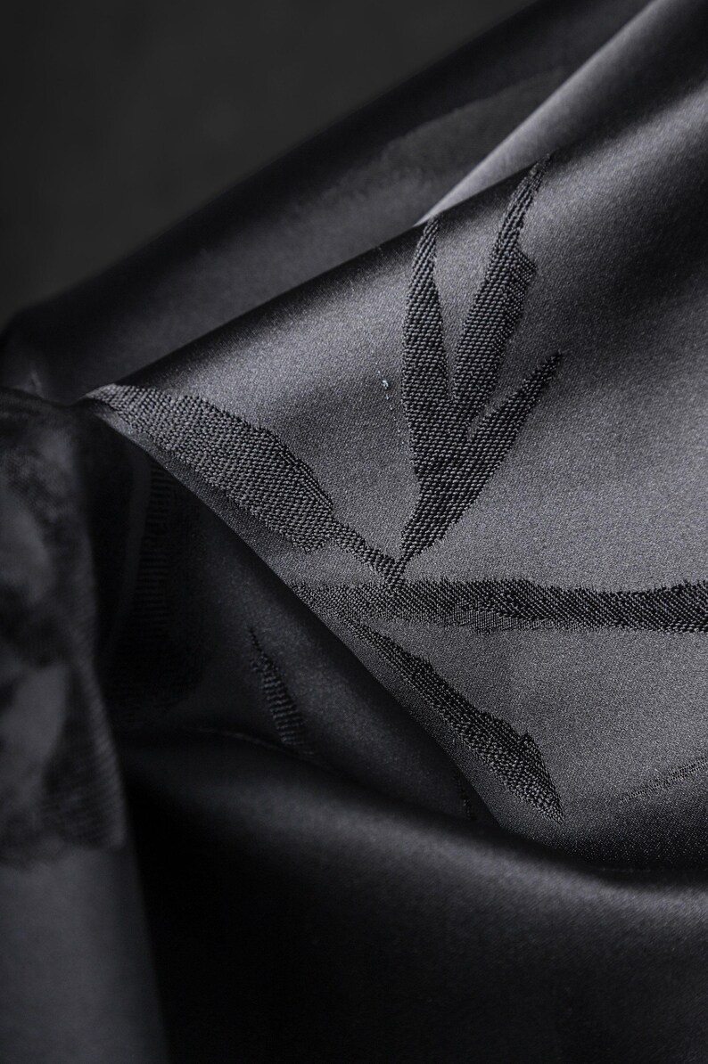 Puede incluir: Primer plano de tela negra con un brillo sutil. La tela est&aacute; doblada, revelando un patr&oacute;n texturizado de hojas o ramas. El color oscuro y el dise&ntilde;o detallado sugieren un textil lujoso, posiblemente para ropa o decoraci&oacute;n del hogar.