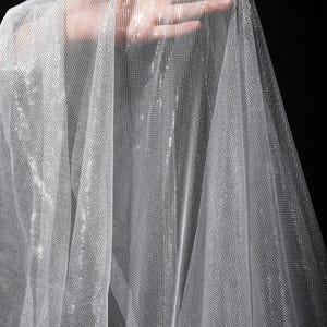 Op de afbeelding: Een doorschijnende, glinsterende stof met een fijn, rasterachtig patroon. De stof is licht zilverkleurig en de textuur is zichtbaar. Het materiaal is gedrapeerd, met een hand zichtbaar door het materiaal. De achtergrond is zwart.