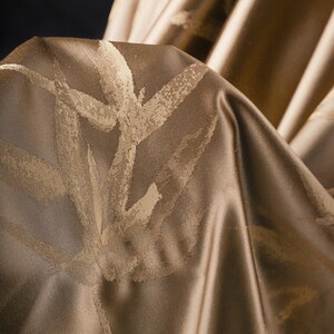 Puede incluir: Primer plano de una tela drapeada en tonos dorados y marrones. El material tiene un ligero brillo y un patr&oacute;n repetido de dise&ntilde;os estilizados de hojas o ramas en un tono dorado m&aacute;s claro. La tela est&aacute; doblada, creando sombras y profundidad.