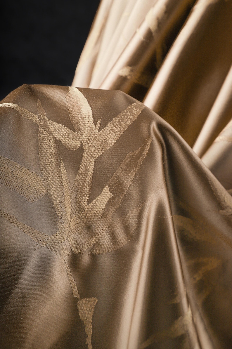 Puede incluir: Primer plano de una tela drapeada en tonos dorados y marrones. El material tiene un ligero brillo y un patr&oacute;n repetido de dise&ntilde;os estilizados de hojas o ramas en un tono dorado m&aacute;s claro. La tela est&aacute; doblada, creando sombras y profundidad.