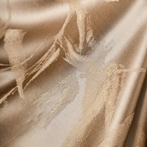 Puede incluir: Primer plano de una tela lujosa y texturizada en un tono beige dorado c&aacute;lido. El material presenta un sutil patr&oacute;n abstracto de pinceladas beige claro, creando una sensaci&oacute;n de profundidad y movimiento. La tela tiene un brillo suave.