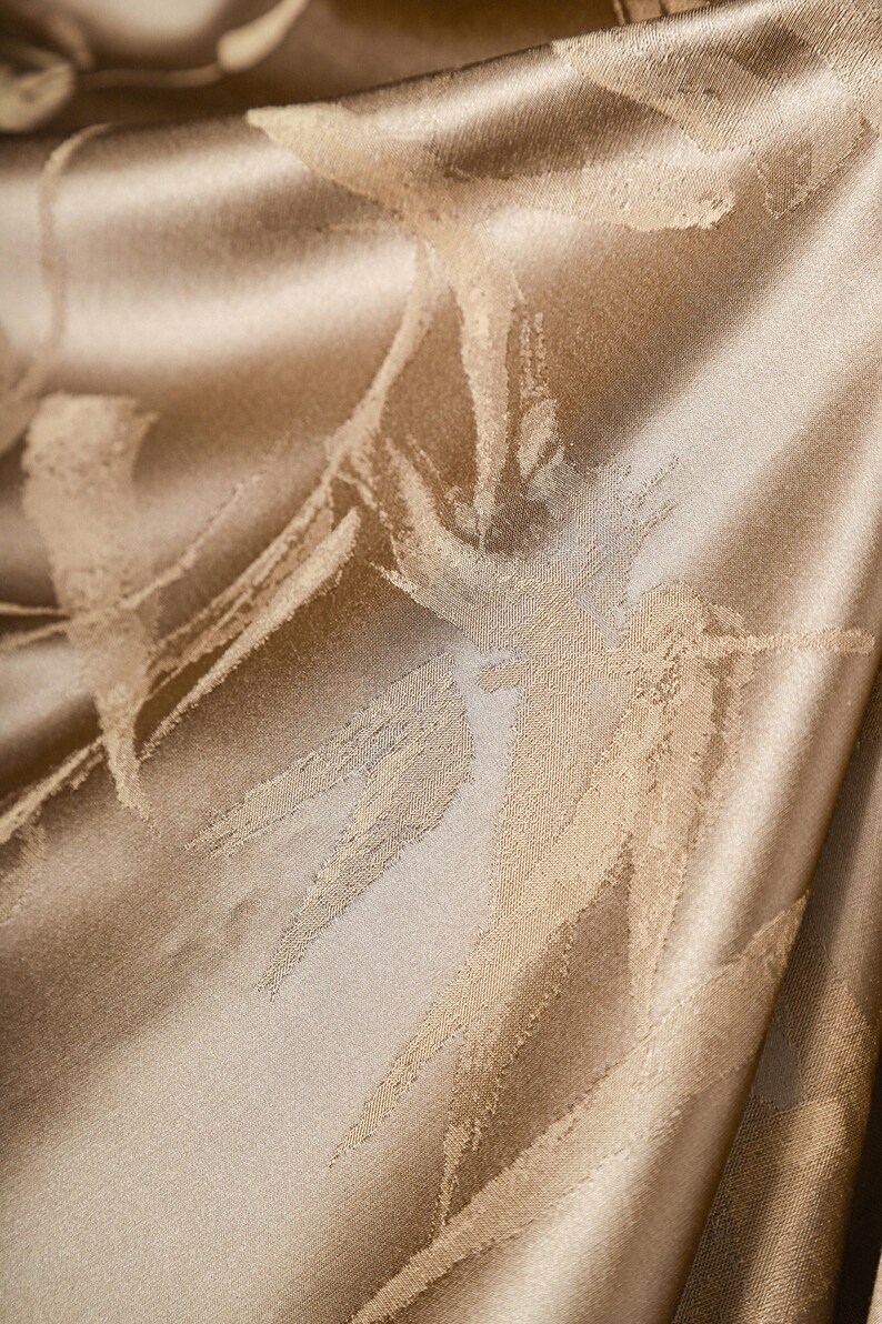 Puede incluir: Primer plano de una tela lujosa y texturizada en un tono beige dorado c&aacute;lido. El material presenta un sutil patr&oacute;n abstracto de pinceladas beige claro, creando una sensaci&oacute;n de profundidad y movimiento. La tela tiene un brillo suave.