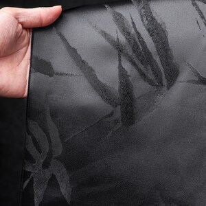 Puede incluir: Tela negra con un sutil estampado de hojas de bamb&uacute;. La tela se sostiene, revelando la textura y el dise&ntilde;o. El color oscuro y el patr&oacute;n sugieren un uso formal o decorativo, posiblemente para ropa o decoraci&oacute;n del hogar.