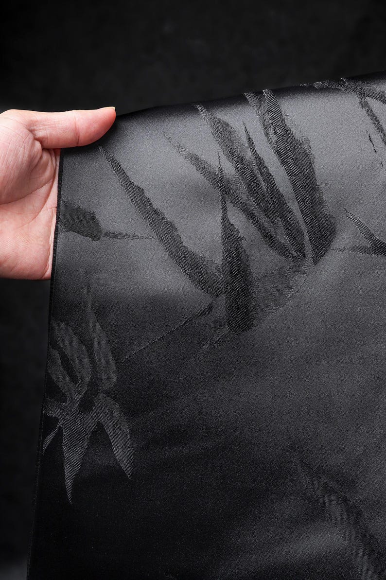 Puede incluir: Tela negra con un sutil estampado de hojas de bamb&uacute;. La tela se sostiene, revelando la textura y el dise&ntilde;o. El color oscuro y el patr&oacute;n sugieren un uso formal o decorativo, posiblemente para ropa o decoraci&oacute;n del hogar.
