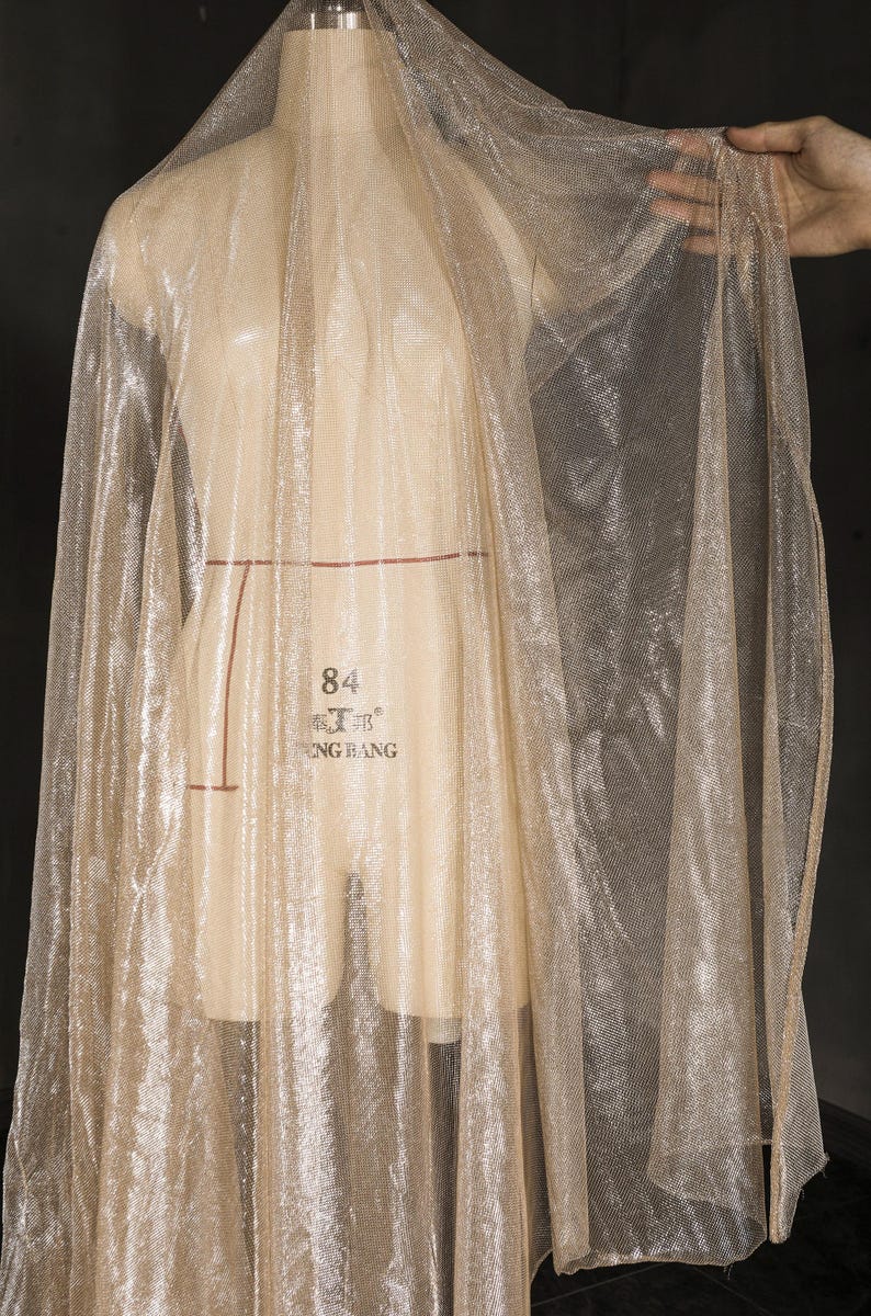 Op de afbeelding: Een doorschijnende, glinsterende stof in een lichtgouden kleur is over een paspop gedrapeerd. De stof heeft een delicate, open weefsel, waardoor een lichte en luchtige uitstraling ontstaat. De paspop is beige en de achtergrond is donker.