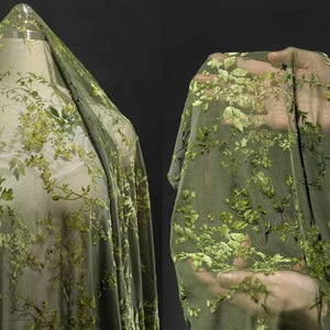 May include: Olive green sheer fabric with embroidered floral and leaf patterns. The fabric is draped over a mannequin and held up by a hand, showcasing the delicate texture and design. The embroidery features varying shades of green.
