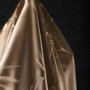 Puede incluir: Una tela drapeada y lustrosa en un tono marr&oacute;n dorado c&aacute;lido. El material presenta un patr&oacute;n sutil y repetitivo de pinceladas abstractas de color claro. Los pliegues de la tela crean sombras, realzando la textura y la profundidad.