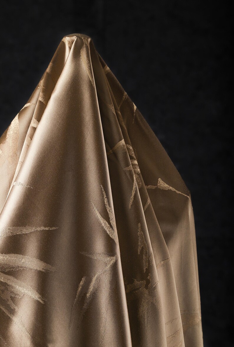 Puede incluir: Una tela drapeada y lustrosa en un tono marr&oacute;n dorado c&aacute;lido. El material presenta un patr&oacute;n sutil y repetitivo de pinceladas abstractas de color claro. Los pliegues de la tela crean sombras, realzando la textura y la profundidad.