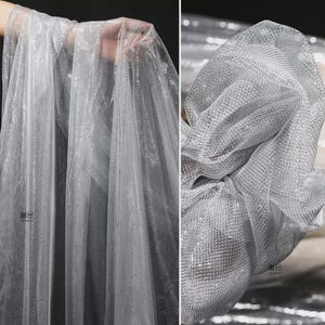 Op de afbeelding: Zilveren glitter tule stof. De stof is transparant en lichtgewicht, met een fijne mesh constructie. De glitter voegt een glinsterend effect toe. De stof wordt gedrapeerd en geplooid getoond, wat de textuur en de val benadrukt.