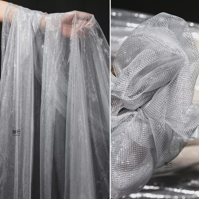 Op de afbeelding: Zilveren glitter tule stof. De stof is transparant en lichtgewicht, met een fijne mesh constructie. De glitter voegt een glinsterend effect toe. De stof wordt gedrapeerd en geplooid getoond, wat de textuur en de val benadrukt.
