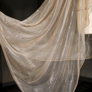 Op de afbeelding: Een vloeiende, transparante stof in een glinsterende goudkleur. Het materiaal is gedrapeerd, waardoor zachte plooien en een delicate textuur ontstaan. De stof lijkt licht van gewicht en kan worden gebruikt voor decoratieve doeleinden of kleding.