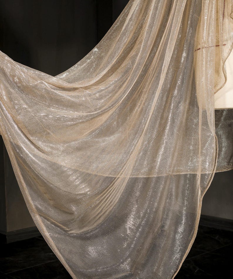 Op de afbeelding: Een vloeiende, transparante stof in een glinsterende goudkleur. Het materiaal is gedrapeerd, waardoor zachte plooien en een delicate textuur ontstaan. De stof lijkt licht van gewicht en kan worden gebruikt voor decoratieve doeleinden of kleding.