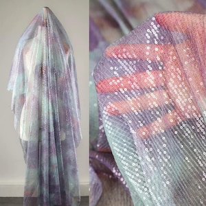 May include: A sheer fabric with a sequined overlay. The fabric is draped over a mannequin, showcasing a watercolor-like pattern in shades of purple, blue, and green. The sequins add a shimmering effect.