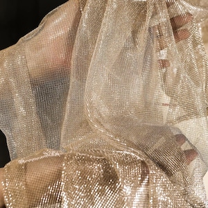 Op de afbeelding: Close-up van een glinsterende gouden mesh stof. De stof is gedrapeerd en gevouwen, waardoor een textuureffect ontstaat. Het materiaal lijkt licht en doorschijnend, met een subtiele glans.
