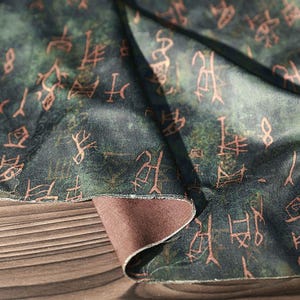 May include: Close-up of a dark green fabric with a brown underside. The fabric is patterned with faded orange-brown symbols. The fabric is draped over a wooden surface, revealing its texture and color variations.
