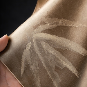 Puede incluir: Primer plano de una secci&oacute;n de tela en un c&aacute;lido tono marr&oacute;n dorado. El material tiene un brillo sutil y presenta un patr&oacute;n repetido de formas de hojas estilizadas en un tono m&aacute;s claro de marr&oacute;n. La tela est&aacute; doblada.