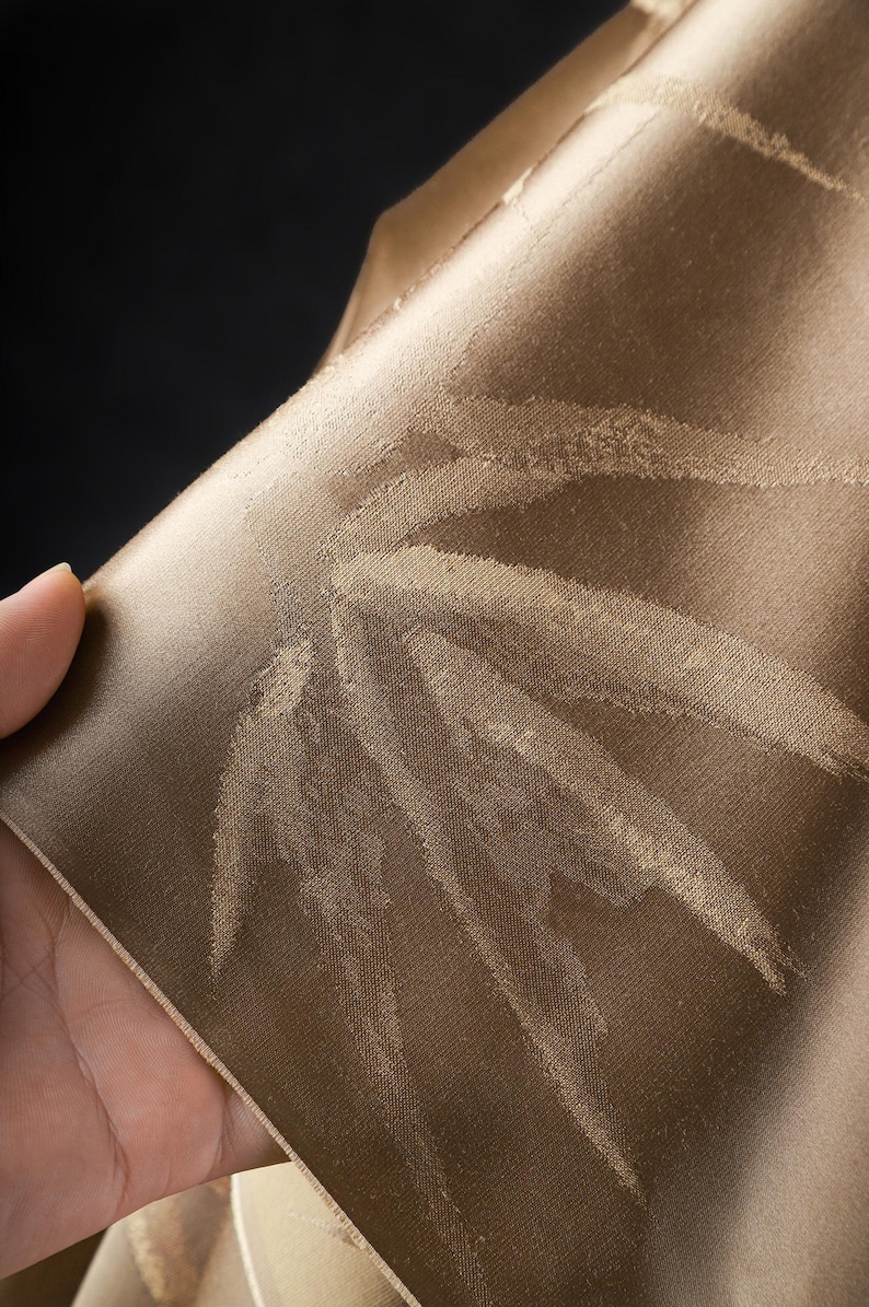 Puede incluir: Primer plano de una secci&oacute;n de tela en un c&aacute;lido tono marr&oacute;n dorado. El material tiene un brillo sutil y presenta un patr&oacute;n repetido de formas de hojas estilizadas en un tono m&aacute;s claro de marr&oacute;n. La tela est&aacute; doblada.