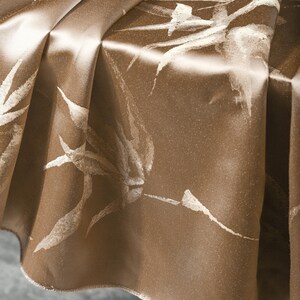 Puede incluir: Primer plano de un mantel de sat&eacute;n marr&oacute;n con un estampado de hojas blancas. La tela est&aacute; drapeada, mostrando pliegues y reflejos. El mantel tiene una textura brillante y suave, adecuado para cenas formales.