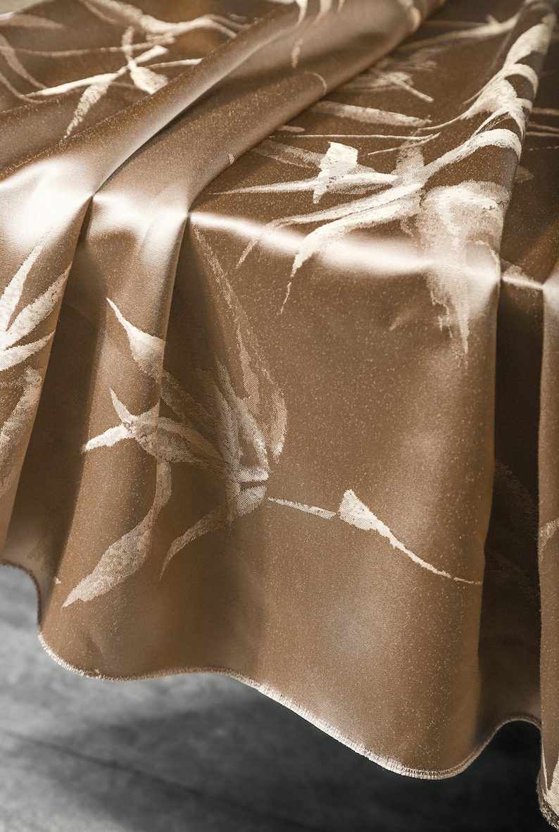 Puede incluir: Primer plano de un mantel de sat&eacute;n marr&oacute;n con un estampado de hojas blancas. La tela est&aacute; drapeada, mostrando pliegues y reflejos. El mantel tiene una textura brillante y suave, adecuado para cenas formales.