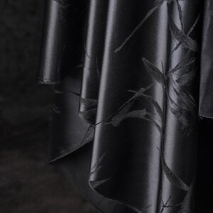 Puede incluir: Primer plano de tela negra con un sutil estampado floral. La tela se pliega en suaves pliegues, creando sombras y reflejos. El material tiene un ligero brillo, realzando el color oscuro y el delicado dise&ntilde;o. El efecto general es elegante y sofisticado.