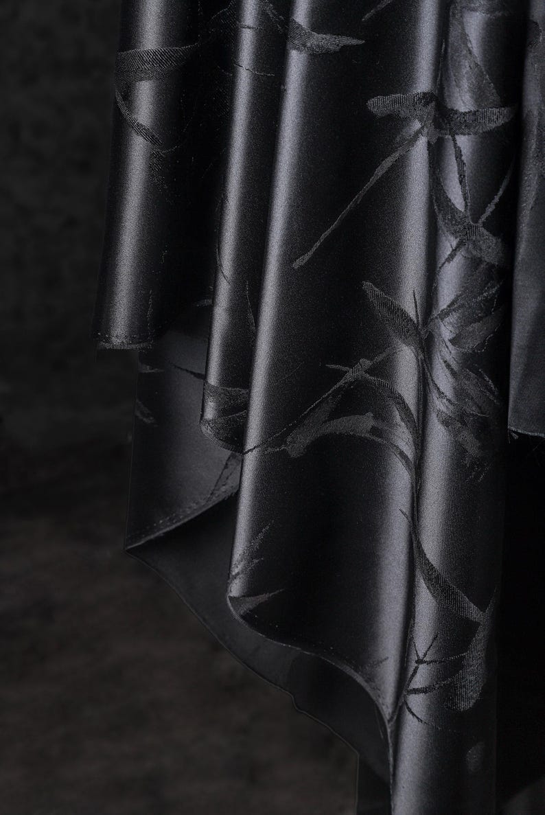 Puede incluir: Primer plano de tela negra con un sutil estampado floral. La tela se pliega en suaves pliegues, creando sombras y reflejos. El material tiene un ligero brillo, realzando el color oscuro y el delicado dise&ntilde;o. El efecto general es elegante y sofisticado.