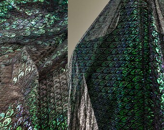 May include: Close-up of a shimmering fabric with a peacock feather pattern. The fabric features a blend of green, black, and silver colors. The material appears to be lightweight and flowing, with a textured surface. Ideal for apparel and craft projects.