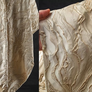 May include: Close-up of a cream-colored fabric with a raised, textured pattern. The design features flowing, organic lines in a lighter gold color, creating a layered effect. The fabric appears to be a luxurious material, possibly for apparel or home decor.