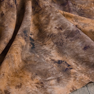 Puede incluir: Primer plano de una tela con un patrón veteado en tonos marrones, beige y negro. La tela tiene un aspecto texturizado con pliegues visibles y variaciones de color, creando una estética natural y orgánica.