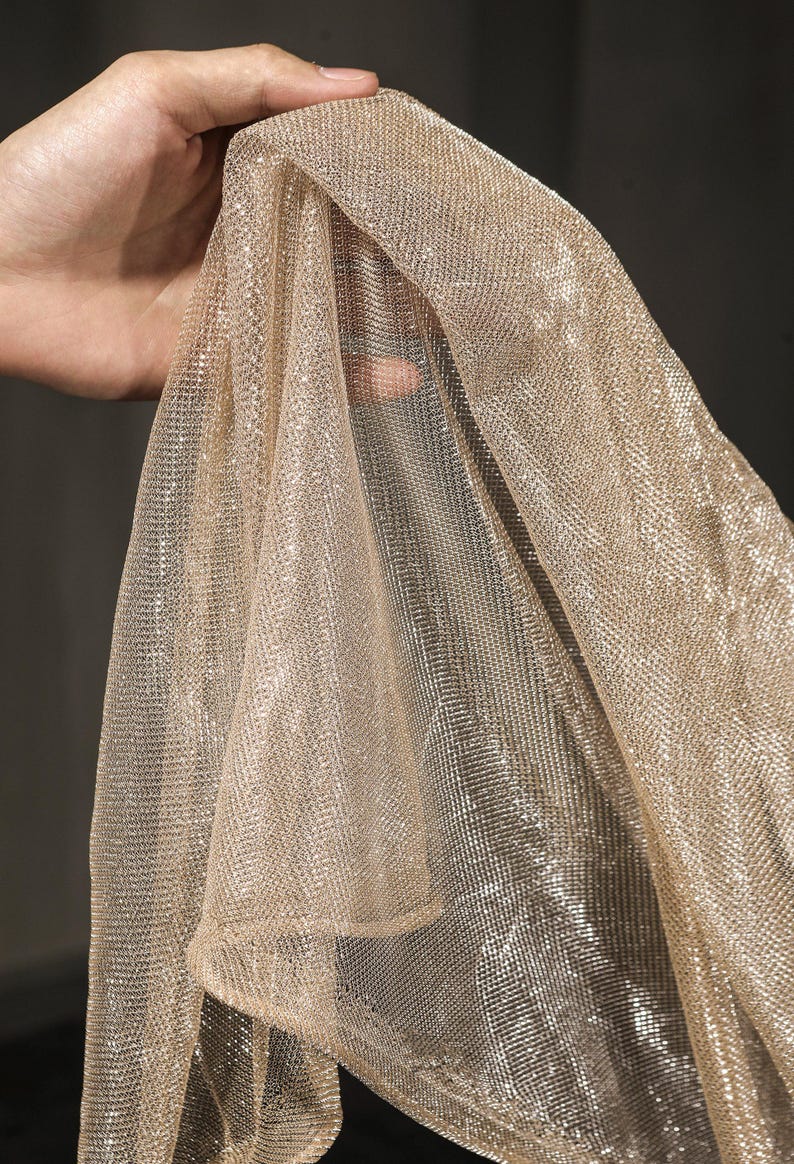 Op de afbeelding: Een glinsterende, goudkleurige stof wordt omhoog gehouden, waarbij de delicate, open weving wordt getoond. Het materiaal heeft een subtiele glans, die het licht vangt en een luxueuze uitstraling cre&euml;ert. De textuur van de stof is zichtbaar, wat duidt op een lichte drapering.
