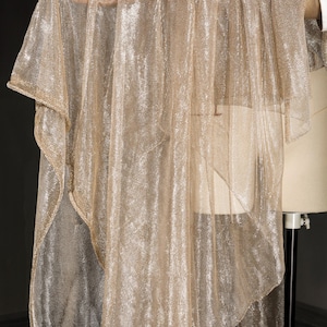Op de afbeelding: Een doorschijnende, goudkleurige stof met een metallic glans is over een mannequin gedrapeerd. De stof heeft een losse weefsel, waardoor een delicate, vloeiende uitstraling ontstaat. Het materiaal wordt waarschijnlijk gebruikt voor kleding of decoratieve doeleinden.