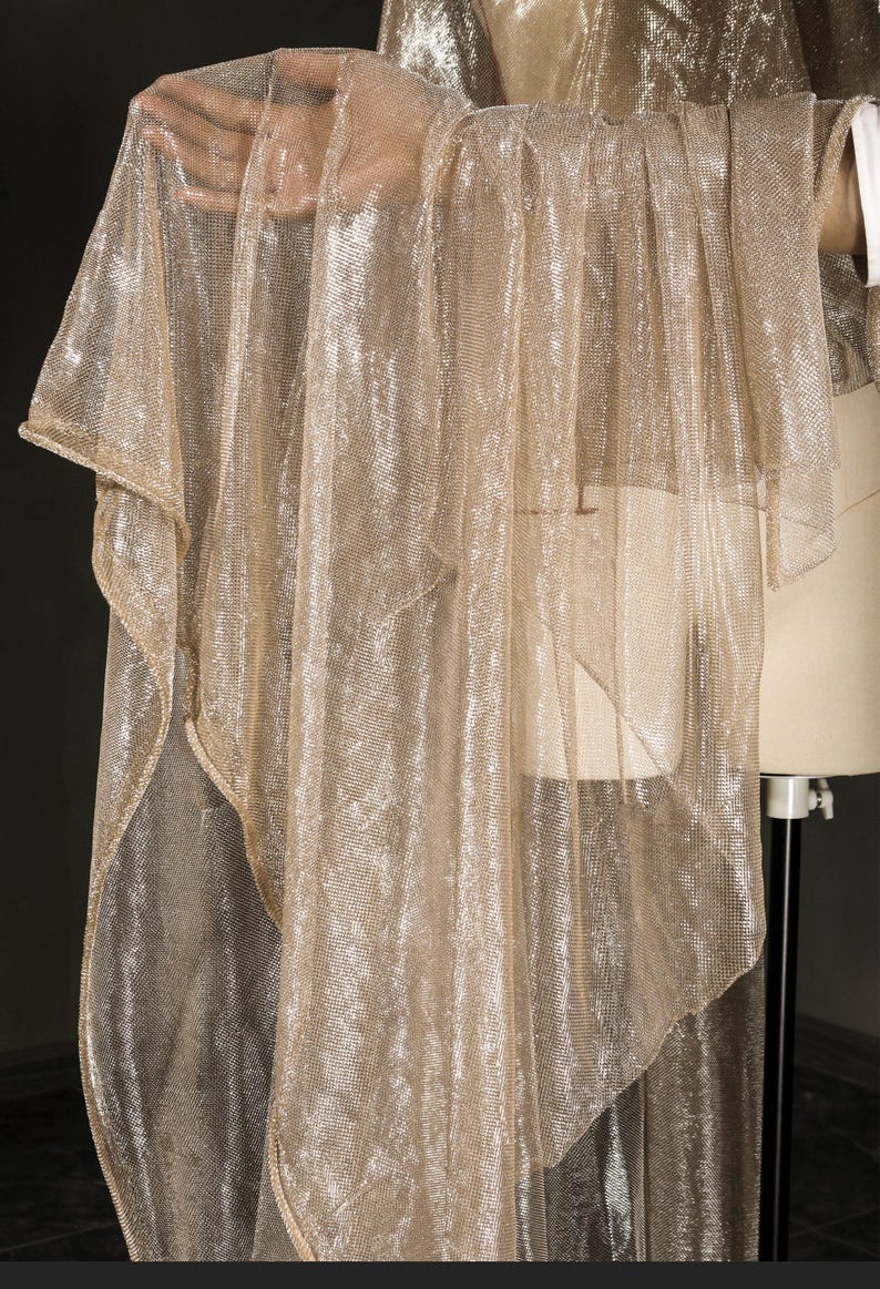 Op de afbeelding: Een doorschijnende, goudkleurige stof met een metallic glans is over een mannequin gedrapeerd. De stof heeft een losse weefsel, waardoor een delicate, vloeiende uitstraling ontstaat. Het materiaal wordt waarschijnlijk gebruikt voor kleding of decoratieve doeleinden.