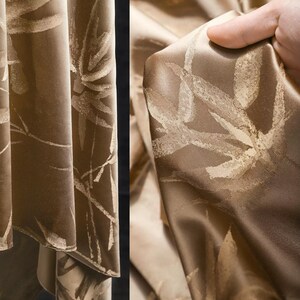 Puede incluir: Primer plano de una tela lujosa drapeada en tonos marrones y beige. La tela presenta un patr&oacute;n repetido de dise&ntilde;os estilizados de hojas o flores de color claro. El material tiene un brillo sutil, lo que sugiere un tejido de seda o sat&eacute;n, y probablemente se utiliza para cortinas o tapicer&iacute;a.