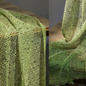 Puede incluir: Primer plano de una tela verde de tejido abierto con aspecto texturizado. La tela tiene un patrón de rejilla y está drapeada sobre una superficie. La tela tiene pequeñas motas de color más oscuro.
