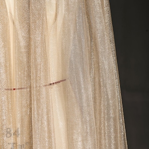 Op de afbeelding: Close-up van een glinsterende, goudkleurige stof met een fijne mesh textuur. Het materiaal drapeert in zachte plooien, vangt het licht op en cre&euml;ert een subtiel, gestructureerd effect. De stof heeft een lichte glans, wat suggereert dat deze voor kleding of decoratieve doeleinden kan worden gebruikt.