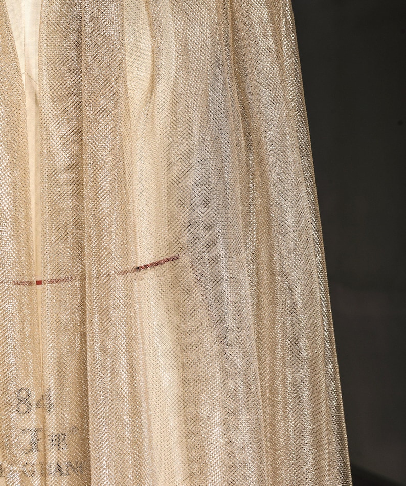 Op de afbeelding: Close-up van een glinsterende, goudkleurige stof met een fijne mesh textuur. Het materiaal drapeert in zachte plooien, vangt het licht op en cre&euml;ert een subtiel, gestructureerd effect. De stof heeft een lichte glans, wat suggereert dat deze voor kleding of decoratieve doeleinden kan worden gebruikt.