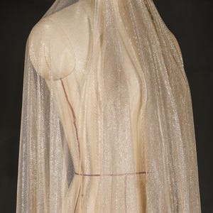 Op de afbeelding: Een doorschijnende, glinsterende stof gedrapeerd over een mannequin. De stof heeft een fijne, geweven textuur en een lichte, gouden tint. De mannequin is neutraal beige van kleur, met rode markeringen. De achtergrond is donker en effen.