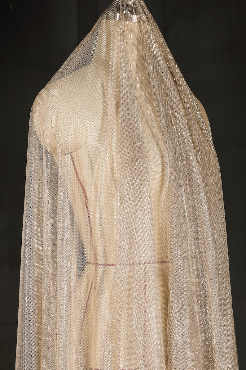 Op de afbeelding: Een doorschijnende, glinsterende stof gedrapeerd over een mannequin. De stof heeft een fijne, geweven textuur en een lichte, gouden tint. De mannequin is neutraal beige van kleur, met rode markeringen. De achtergrond is donker en effen.