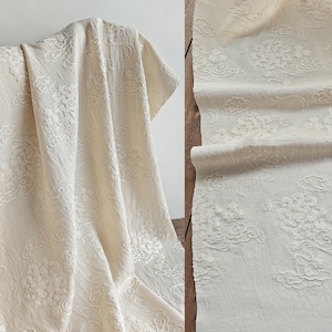 Può includere: Tessuto bianco sporco con un intricato motivo floreale e di nuvole. Il tessuto è drappeggiato su una struttura in legno e srotolato, mostrando il design dettagliato. La trama appare morbida e il colore è un neutro crema.